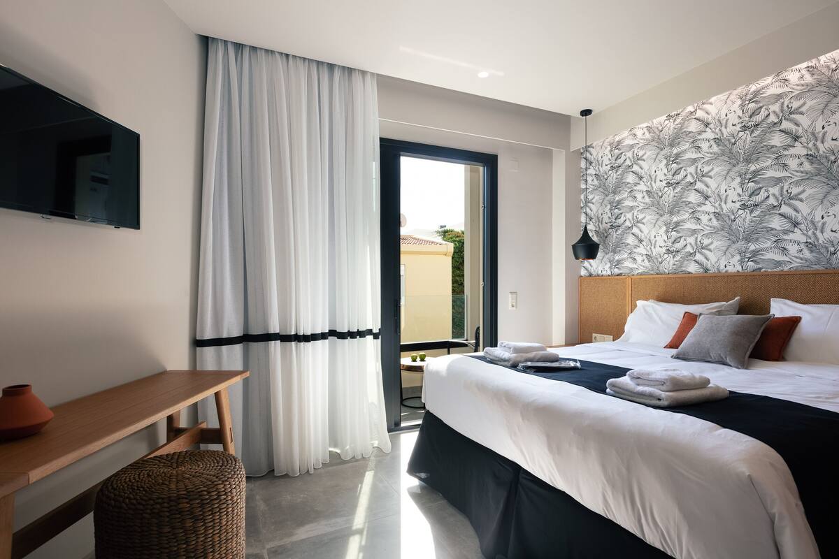 A modern hotel room with a large bed, a flat-screen TV, a wooden desk, and a large window with curtains.