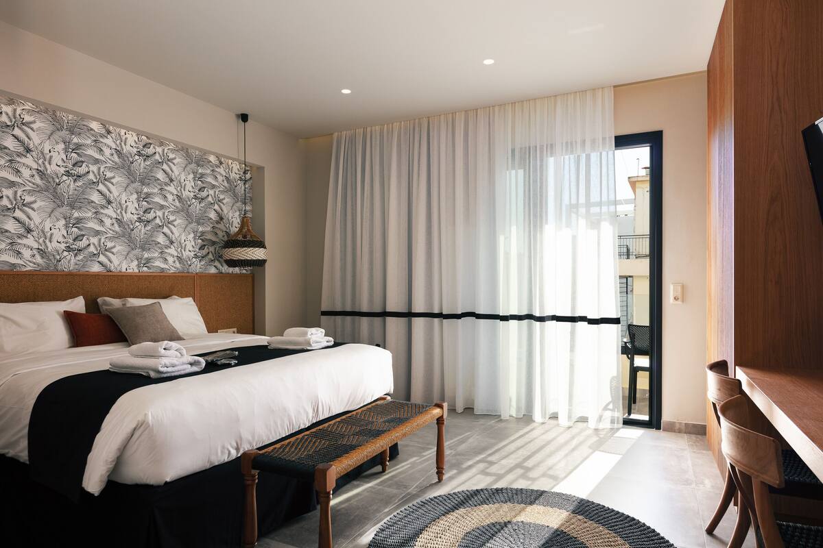 A hotel room with a large bed, a bench, a desk, and a view of the outside.