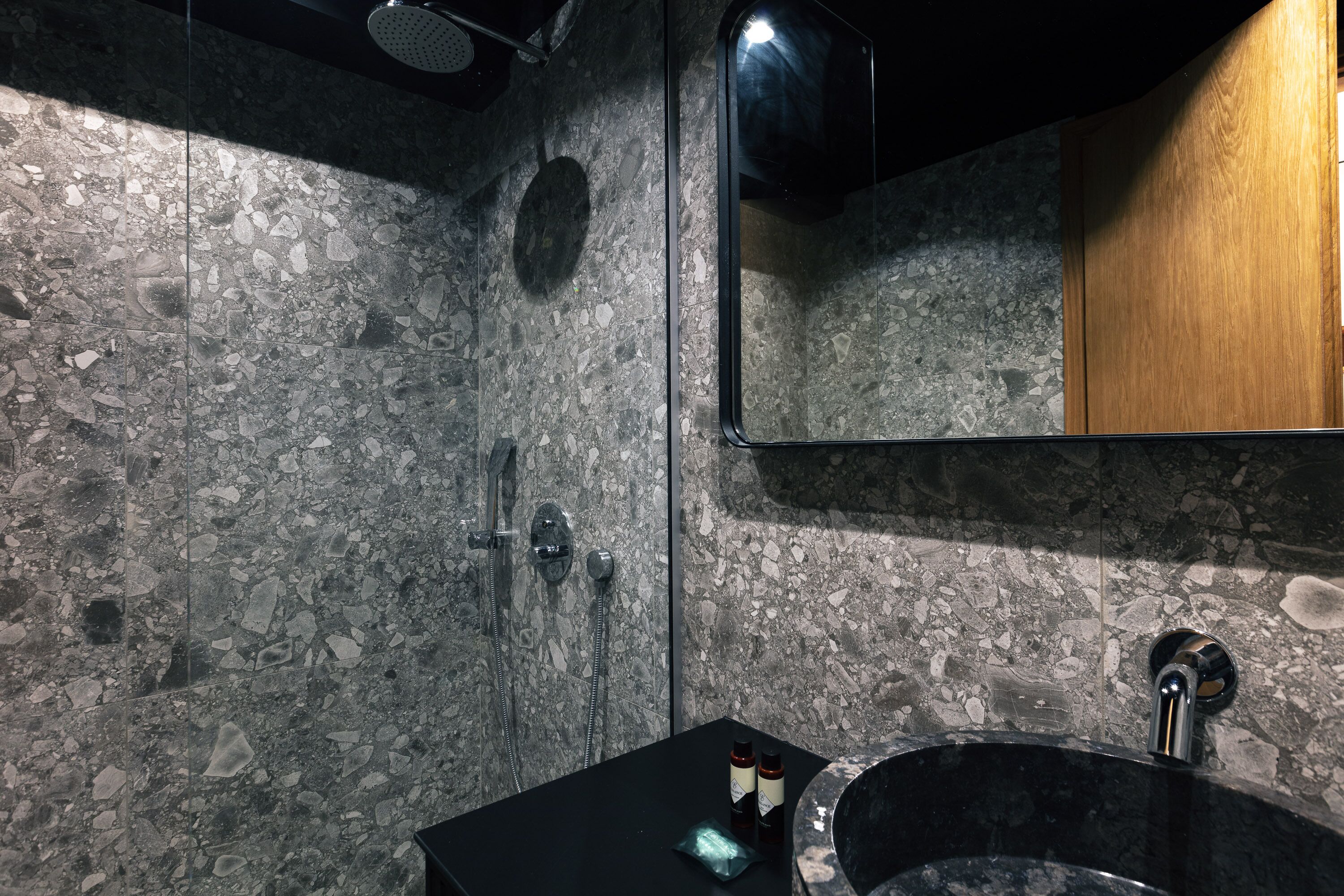 Un baño con pared de granito, un lavabo negro, un espejo y un cabezal de ducha.