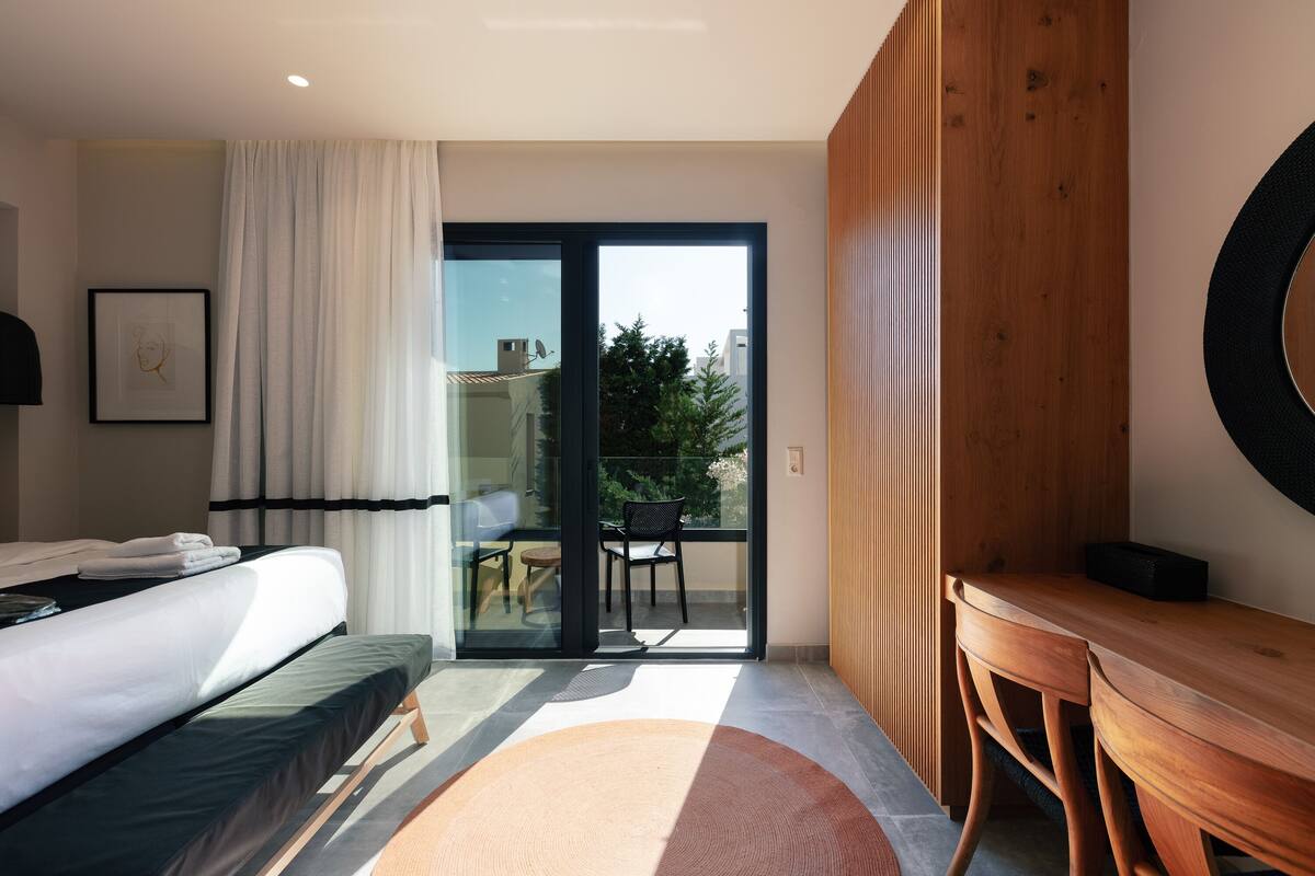 A modern hotel room with a bed, a wooden side table, a large window with curtains, and a view of the outdoors.