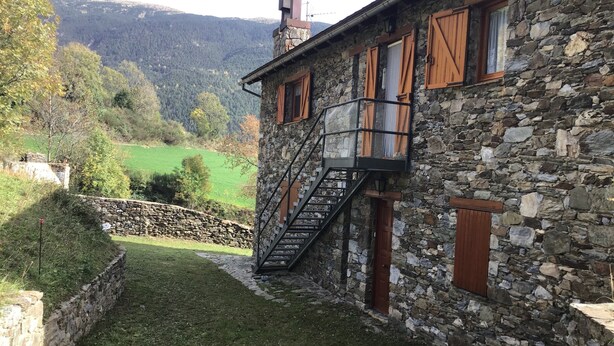 A stone building with a staircase leading to a wooden door, surrounded by a stone wall and a grassy area.