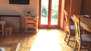 A room with a wooden floor, a TV mounted on the wall, a desk with a chair, and a dining table with chairs.