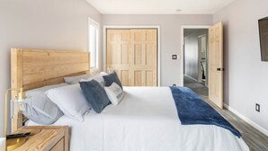 Un dormitorio con cabecera de madera, cama con ropa de cama blanca y gris, manta azul, mesita de noche con lámpara y ventilador de techo.