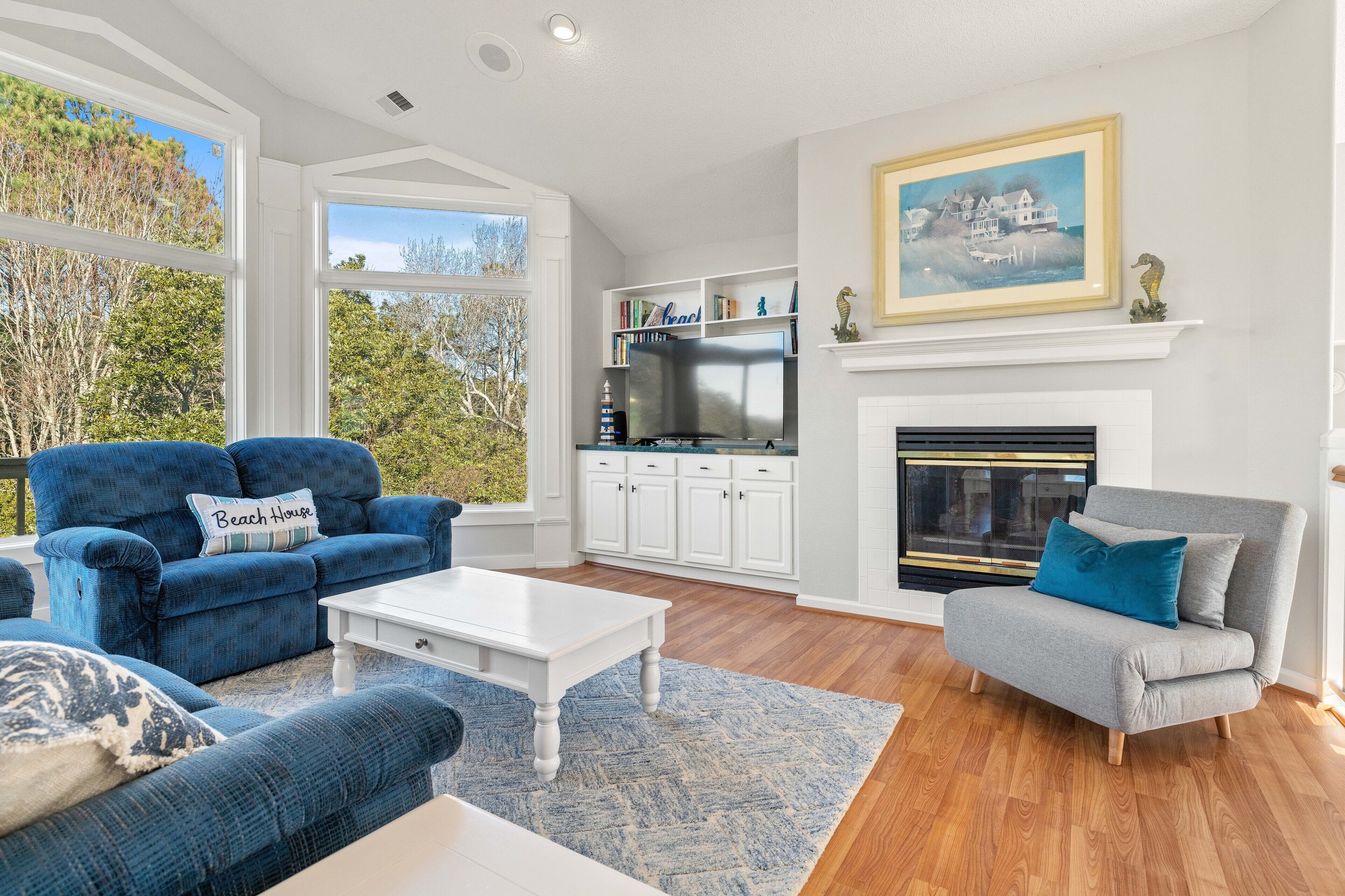 A living room with a fireplace, a television, and a view of trees through large windows.