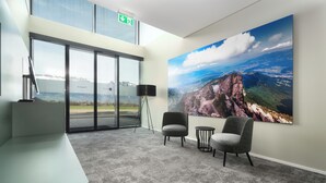 Un espacio de oficina moderno con vistas a un paisaje montañoso a través de amplios ventanales, un mostrador de recepción, dos sillones y una lámpara de pie.