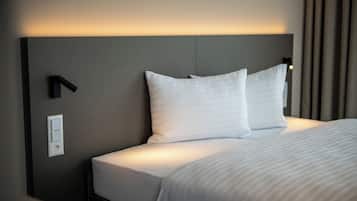 A neatly made bed with white linens and pillows, a wall-mounted light fixture, and a power outlet on the wall.