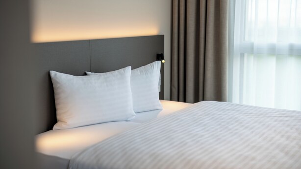 Habitación de hotel moderna con una cama grande, cabecera con iluminación integrada y vistas al exterior a través de cortinas translúcidas.