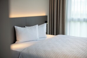 Habitación de hotel moderna con una cama grande, cabecera con iluminación integrada y vistas al exterior a través de cortinas translúcidas.