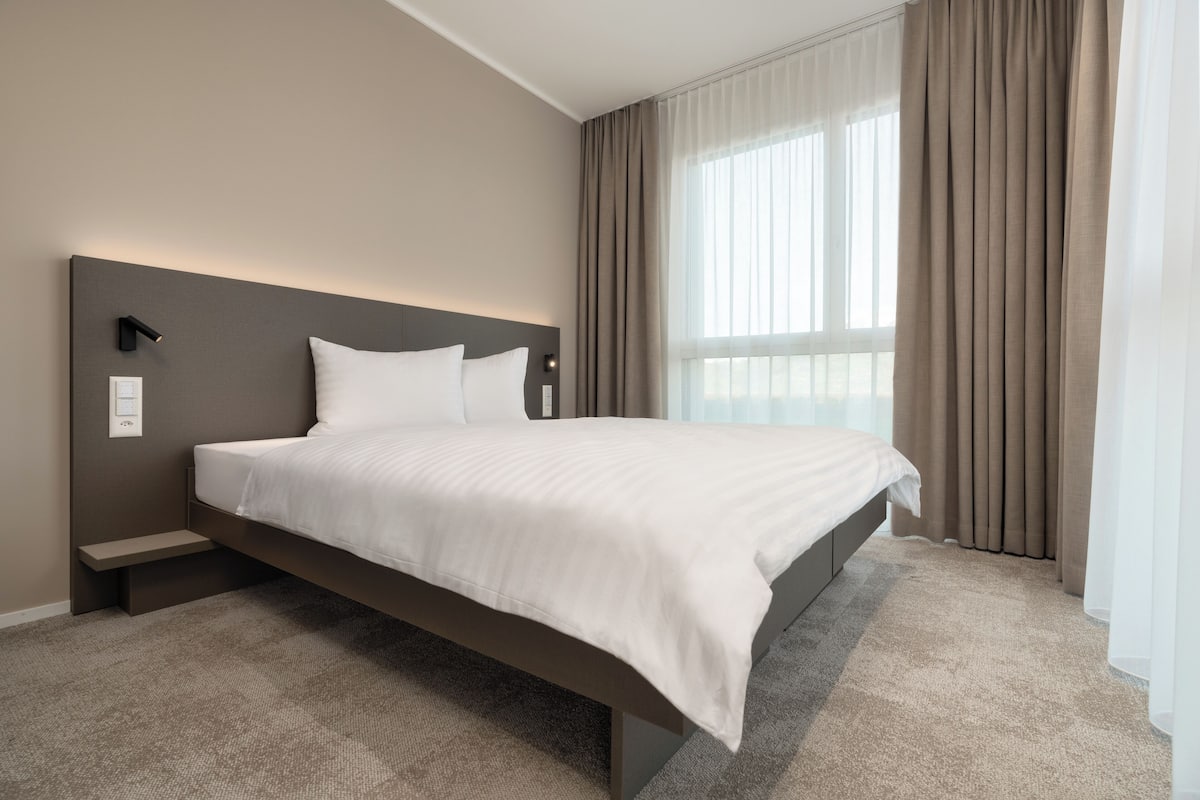 A modern hotel room with a large bed, a bedside table, a wall-mounted light, and a window with curtains.