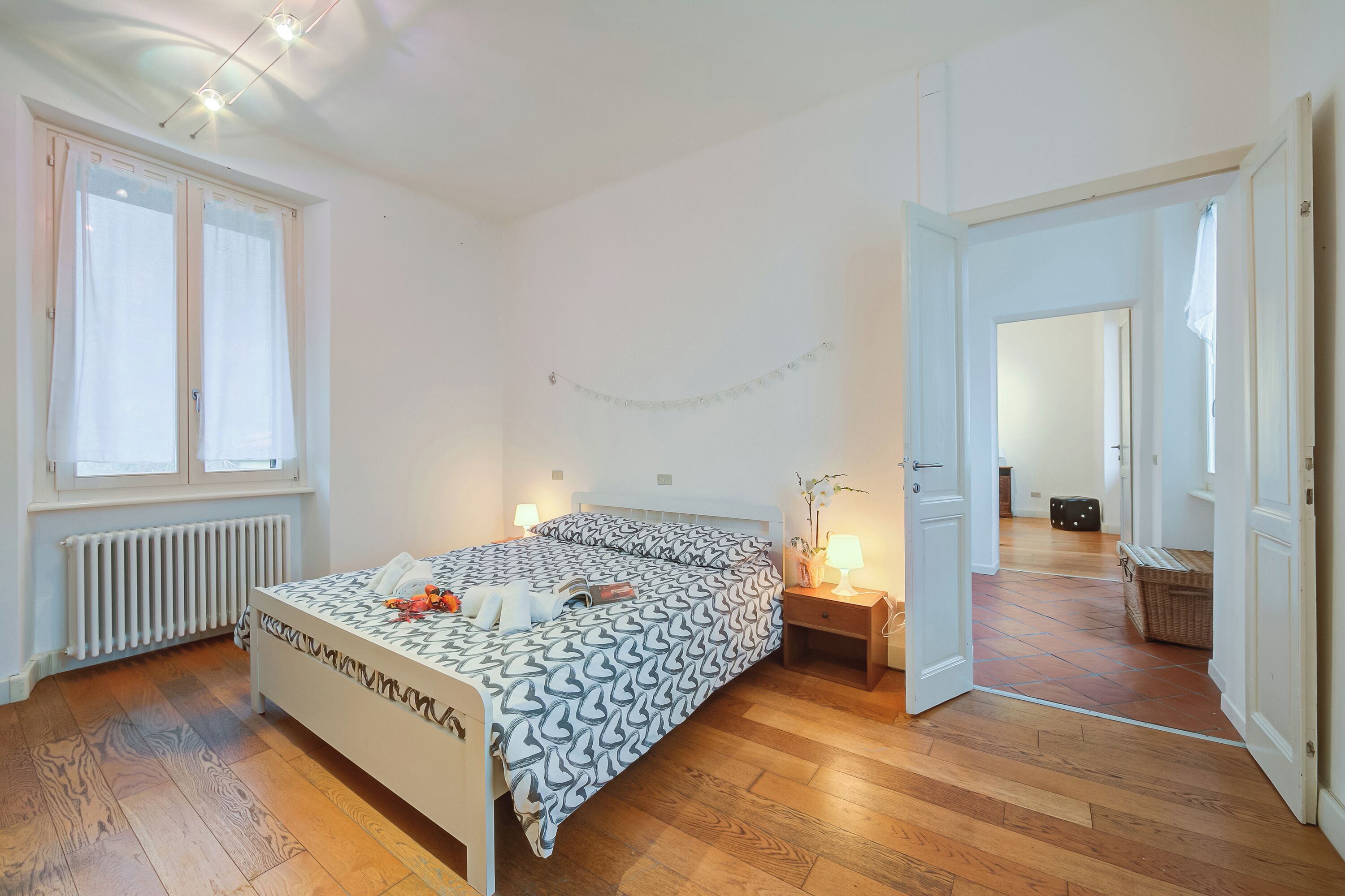 A bedroom with a bed, bedside table, lamp, radiator, window, and wooden floor.