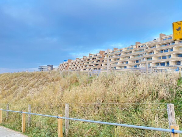 Ein Wohngebäude an der Küste mit mehreren Balkonen, ein sandiger Vordergrund mit Gras und ein klarer Himmel.