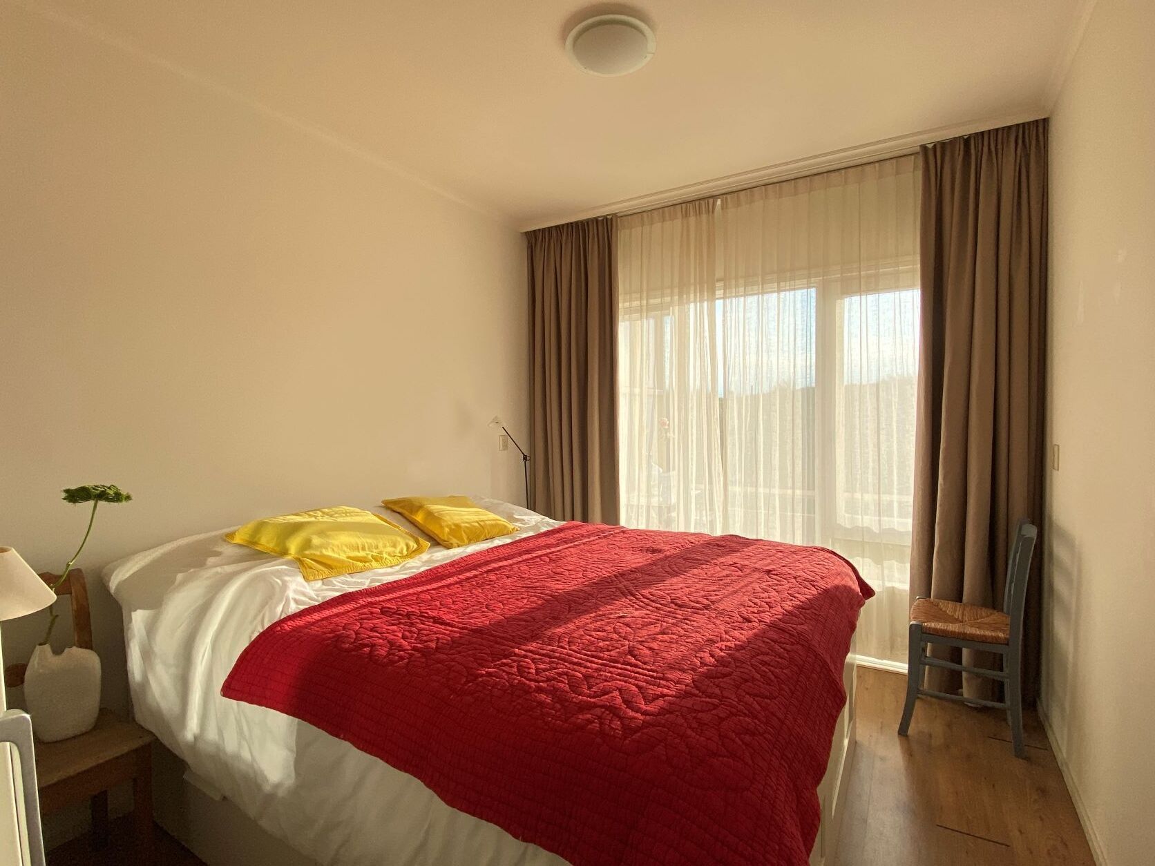 Une chambre d’hôtel avec un lit, une chaise, une lampe et une fenêtre avec des rideaux.