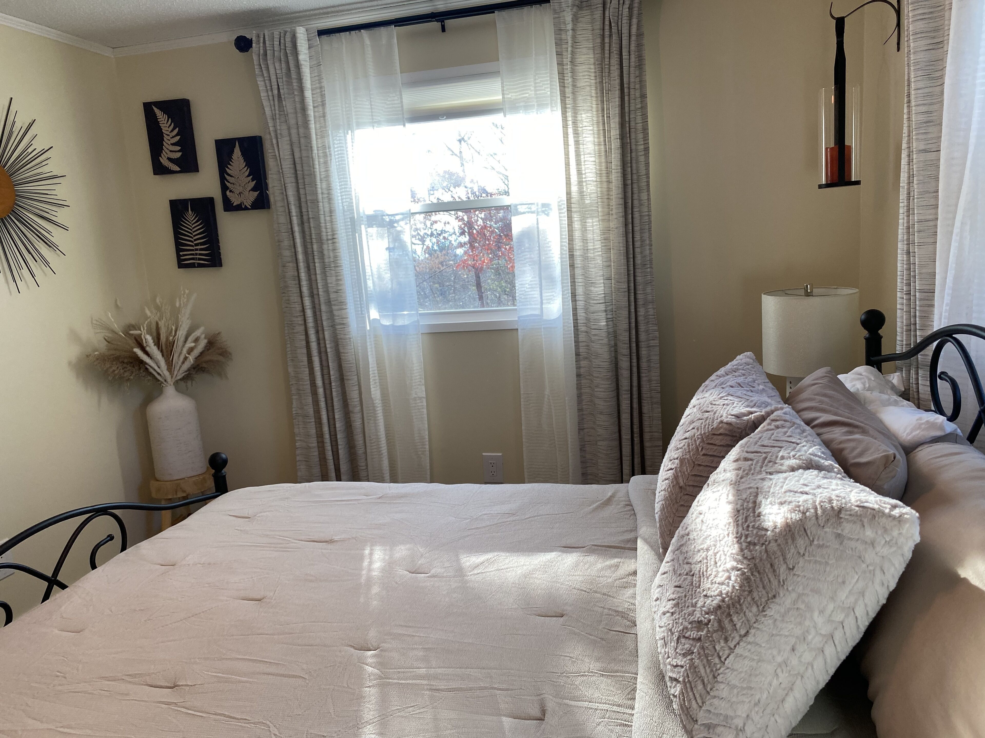 A neatly made bed with a white comforter and pillows, a bedside lamp, a vase with dried plants, and a window with sheer curtains.