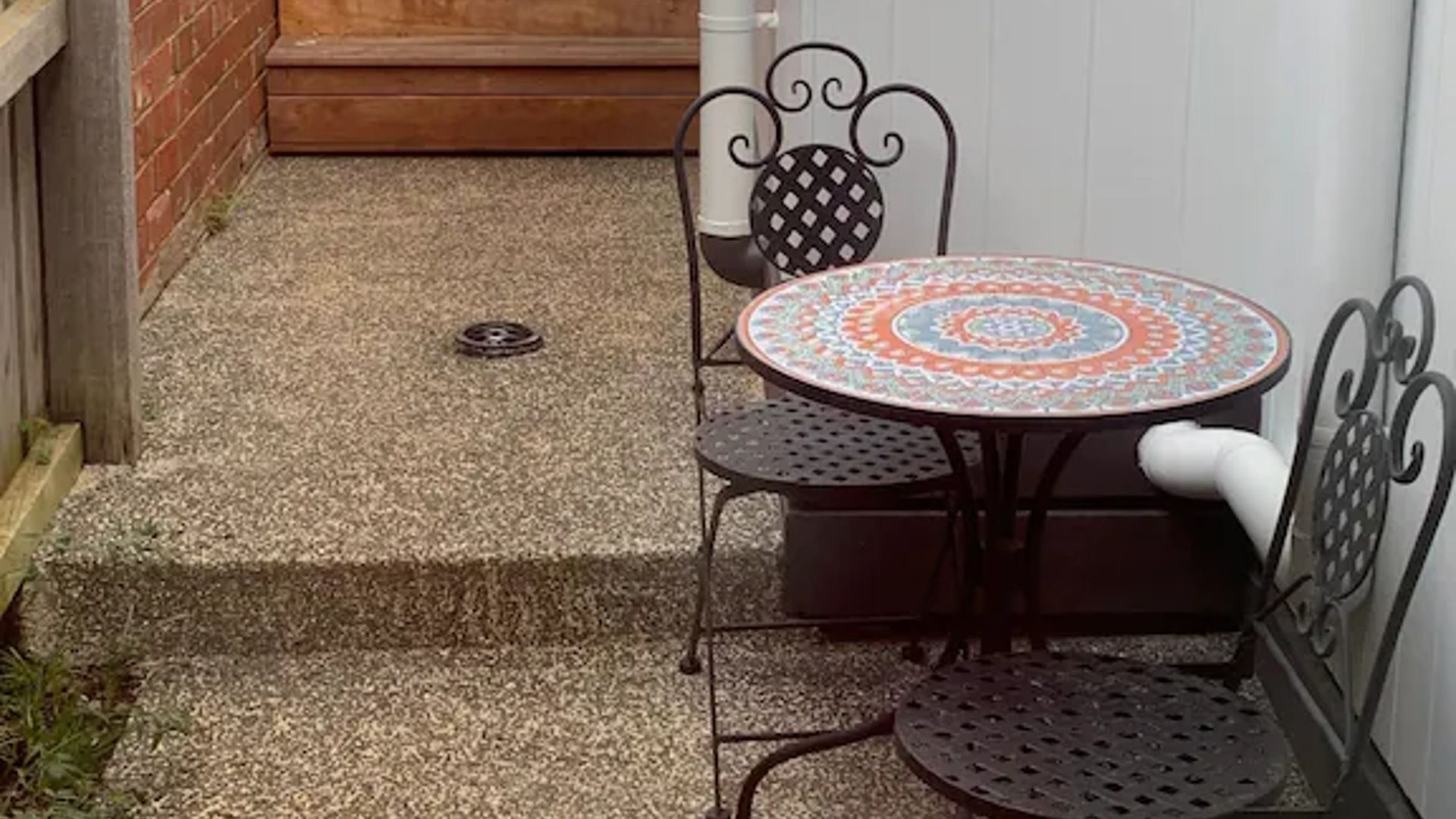 A small patio area with a round table and four chairs, a metal bench, and a manhole cover on the ground.