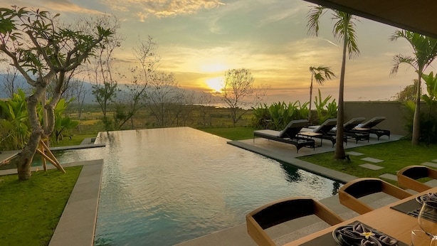 A poolside dining area with a view of the sunset, lounge chairs, and a table set for a meal. - Villa with sunset sea view and jacuzzi - 700m from the beach! (Buleleng)