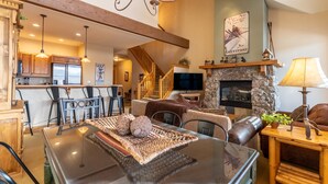 A spacious living area with a dining table, leather sofas, a stone fireplace, and a kitchen in the background. - Ski-in/Ski-out Condo Next to Golf Course, Amazing Mountain Views from Balcony (Keystone)