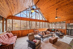A spacious wooden-paneled room with a large window, a ceiling fan, and a cozy seating area with a checkered sofa and wicker chairs.