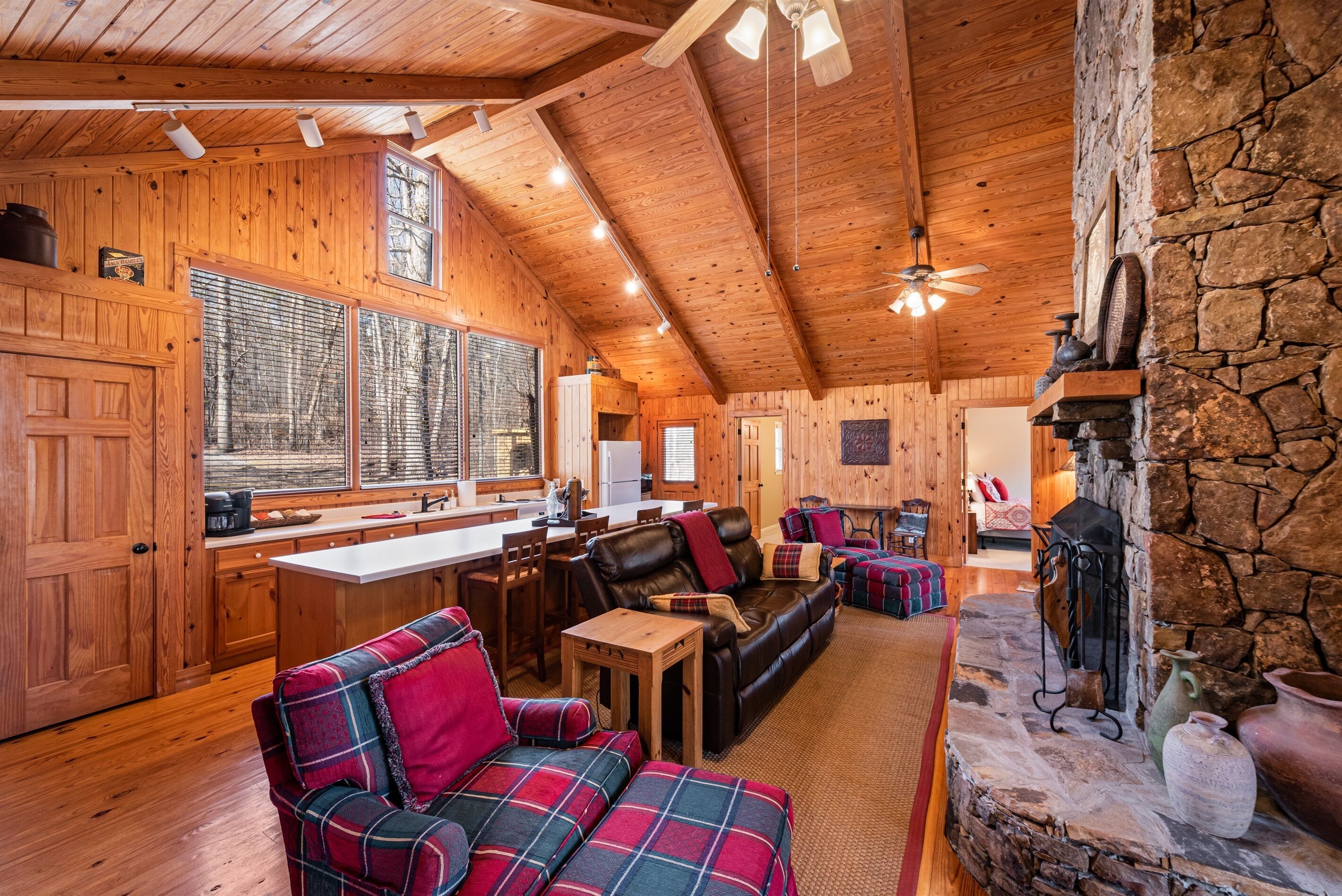 A cozy cabin interior with wooden walls, a stone fireplace, a dining area, and a living room with plaid furniture.
