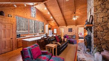A cozy cabin interior with wooden walls, a stone fireplace, a dining area, and a living room with plaid furniture.