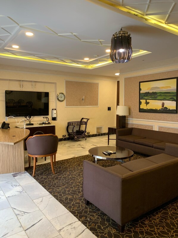 A modern living room with a sofa, coffee table, and TV. There is a desk with a chair, a clock, and a painting on the wall. - BEST PREMIER MAITAMA RESIDENCE (Abuja)