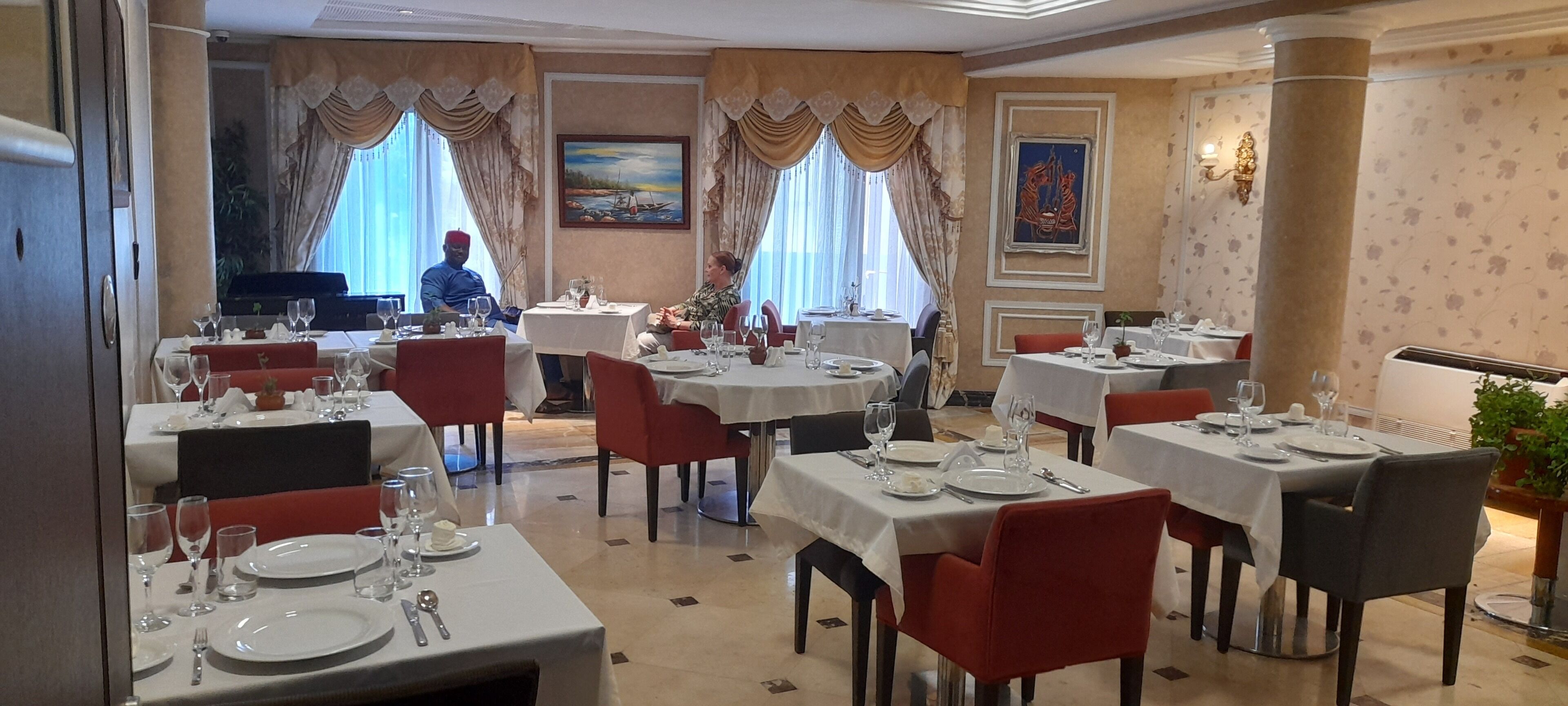 A restaurant with multiple tables, red chairs, and a piano in the background.