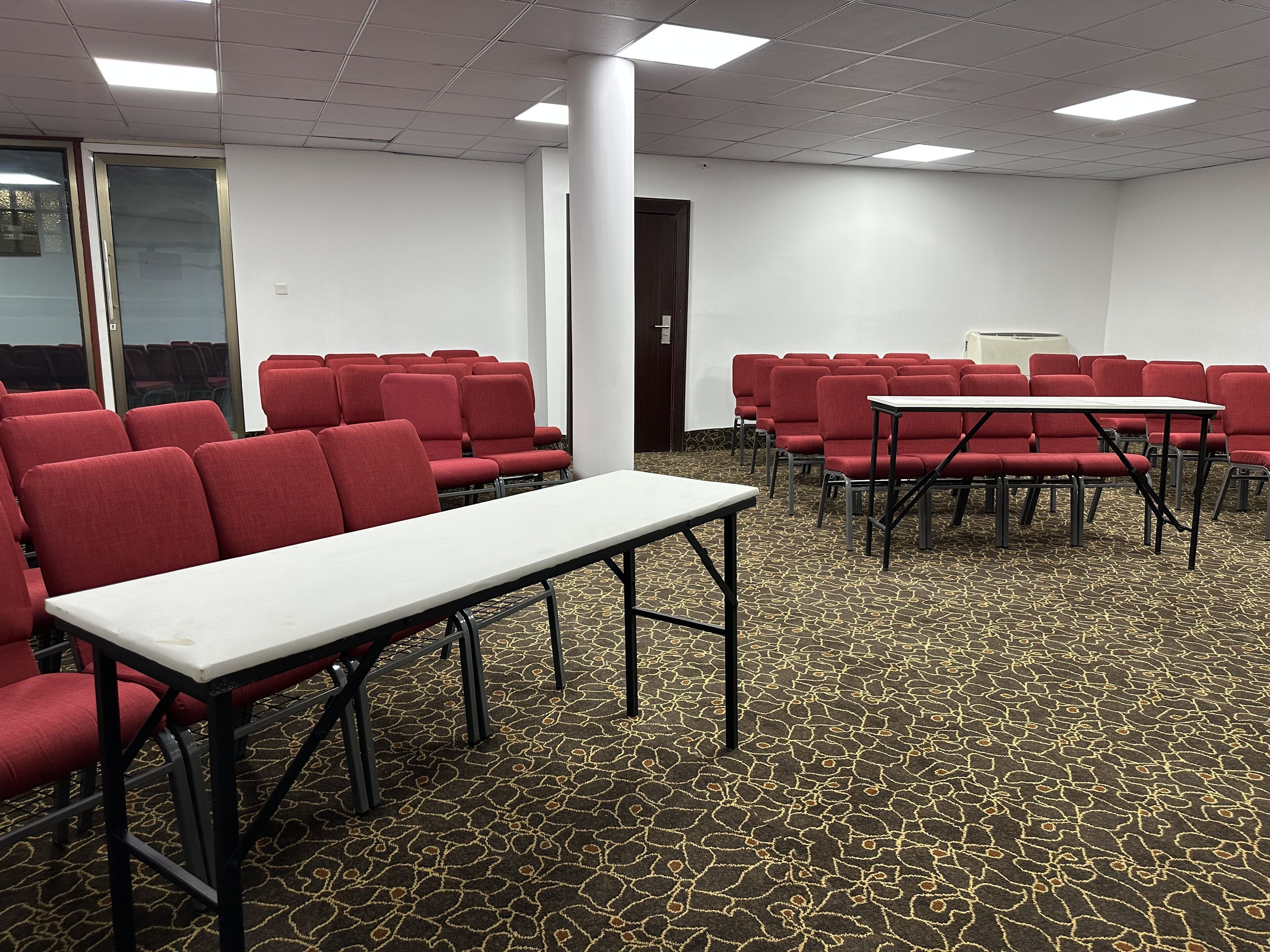 Une salle de conférence avec des rangées de chaises rouges, une table blanche et un tapis à motifs.