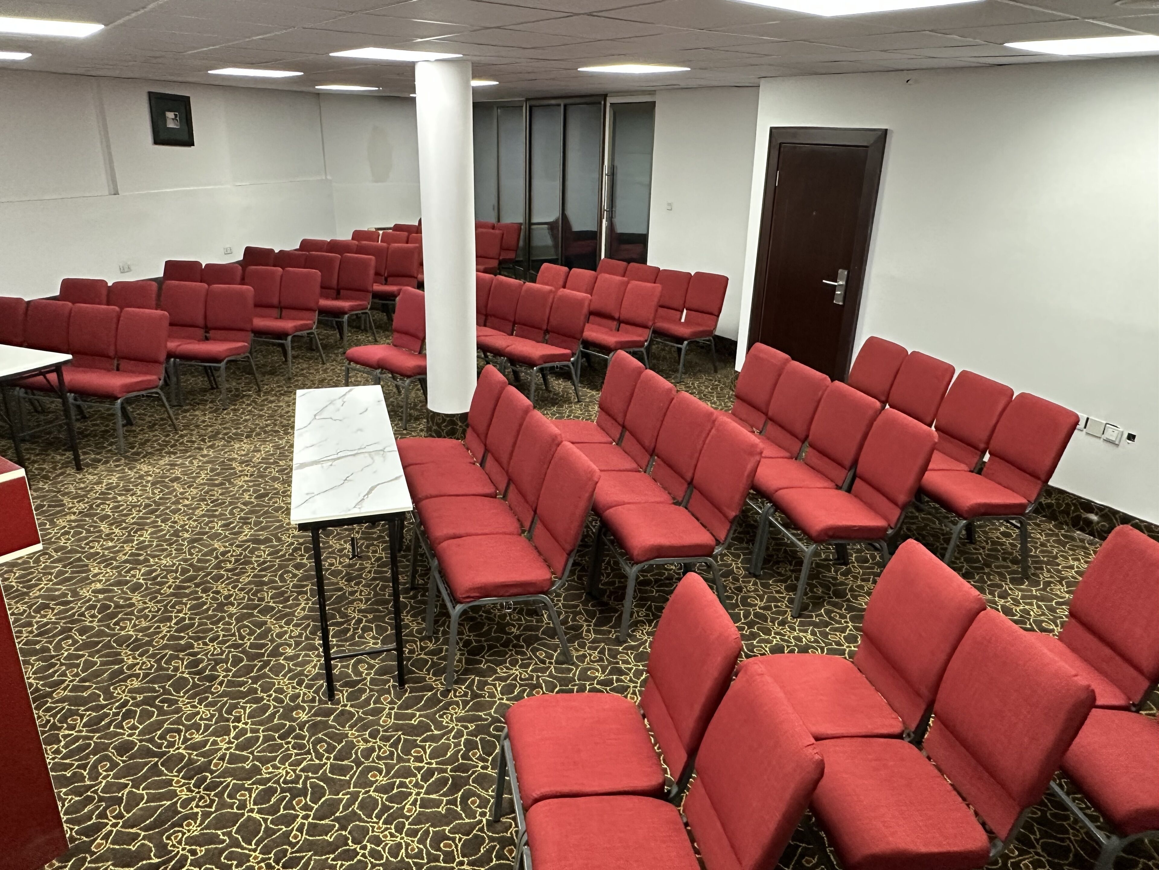 A conference room with rows of red chairs, a white table, and patterned carpet.