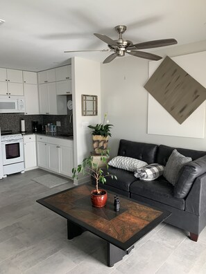 A modern kitchen with white cabinets, a microwave, and a coffee maker. There is a black leather sofa with a gray blanket and pillows. A wooden coffee table with a potted plant is in the center. - Sea Shells- Ocean/Beach Views for miles. (Myrtle Beach)