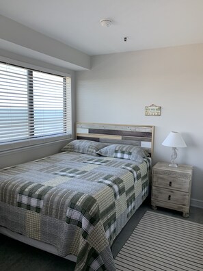 A bedroom with a bed, a nightstand, a lamp, and a window with blinds. - Sea Shells- Ocean/Beach Views for miles. (Myrtle Beach)