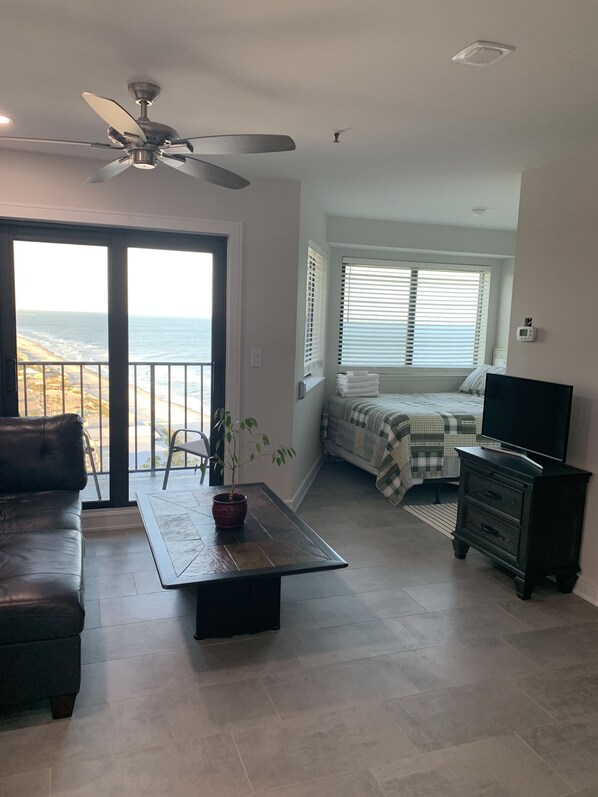 A living room with a sofa, a coffee table, a TV stand, and a ceiling fan. There is a window with blinds and a view of the beach. - Sea Shells- Ocean/Beach Views for miles. (Myrtle Beach)