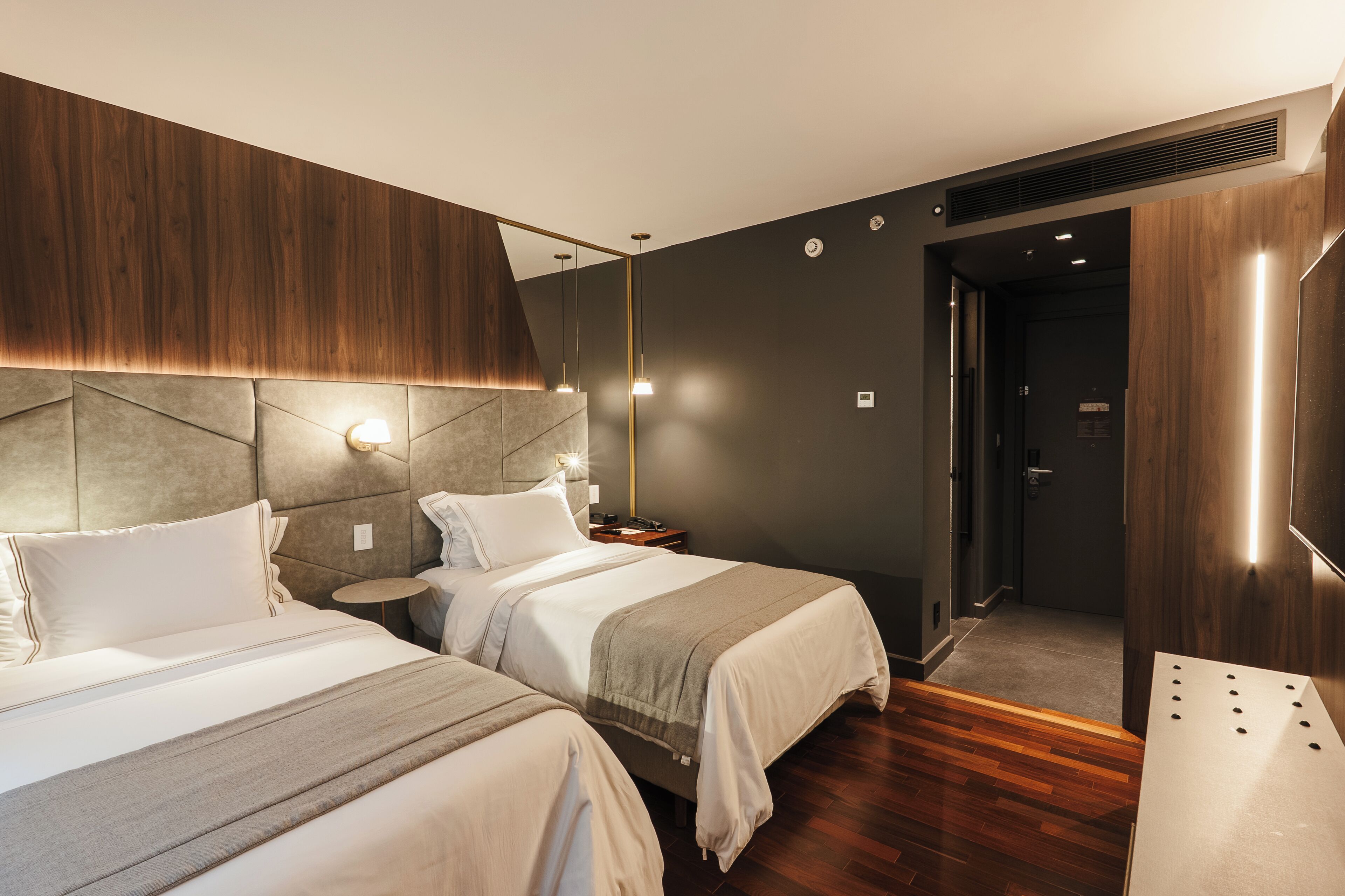 A modern hotel room with two beds, a wooden accent wall, and a small bedside table with a lamp.