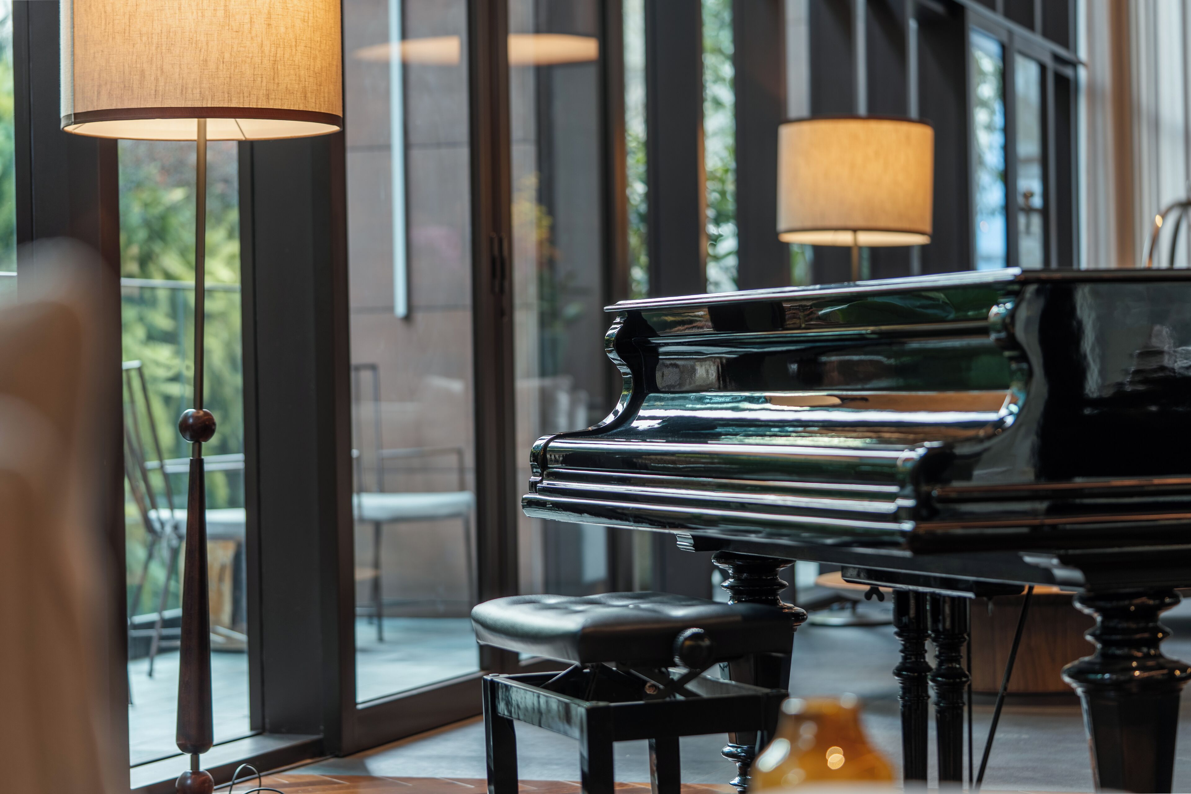 A grand piano in a well-lit room with large windows and floor lamps.