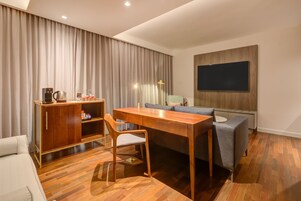 Habitación de hotel moderna con una mesa de comedor de madera, un sofá, un televisor montado en la pared y un minibar.