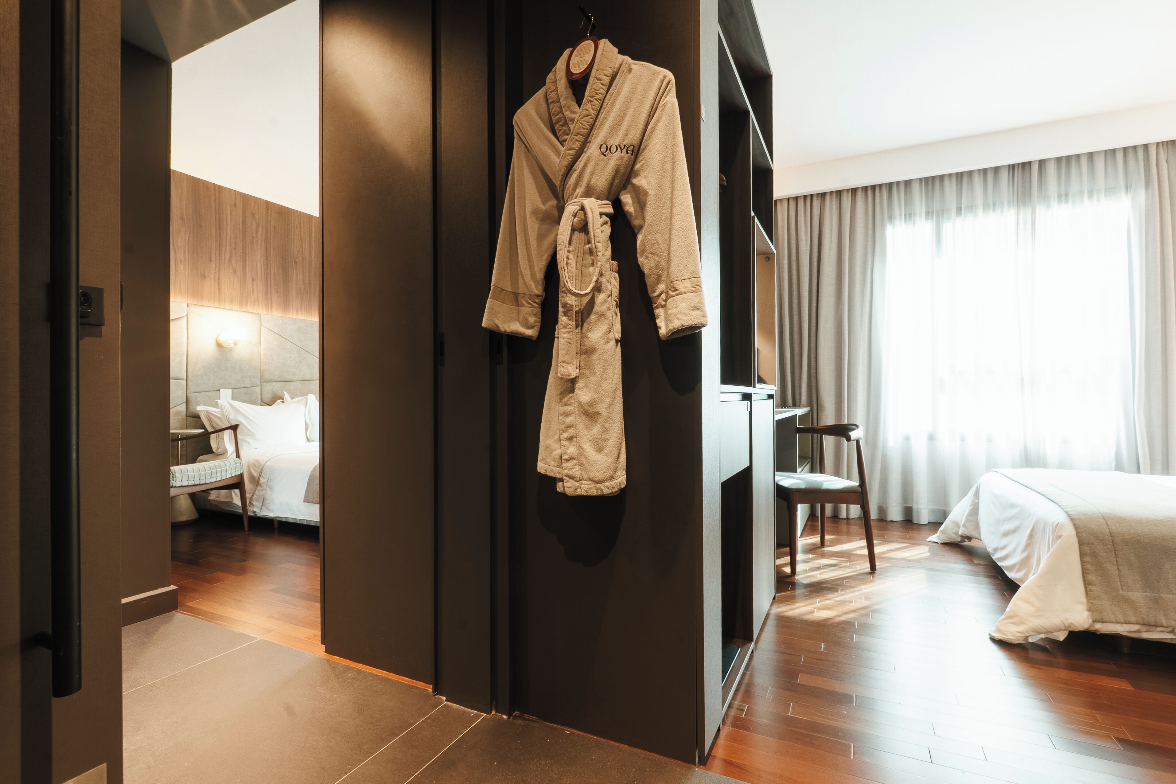 Habitación de hotel con piso de madera, una cama con ropa blanca, una silla, un escritorio y una bata colgada en la pared.