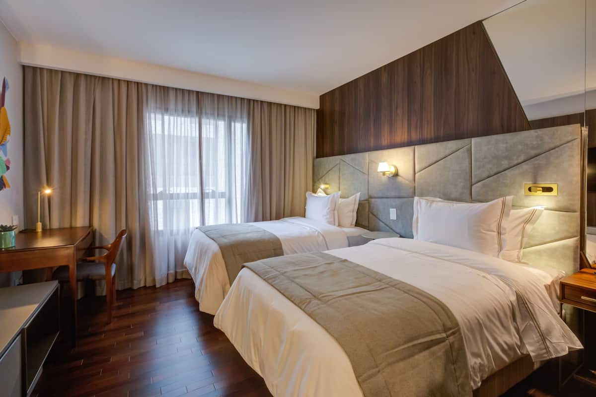Ein Hotelzimmer mit zwei Betten, einem Schreibtisch, einem Stuhl, einem großen Fenster mit Vorhängen und einem Holzkopfende.