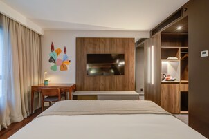 A hotel room with a bed, a desk, a chair, a TV mounted on a wooden panel, and a wardrobe.