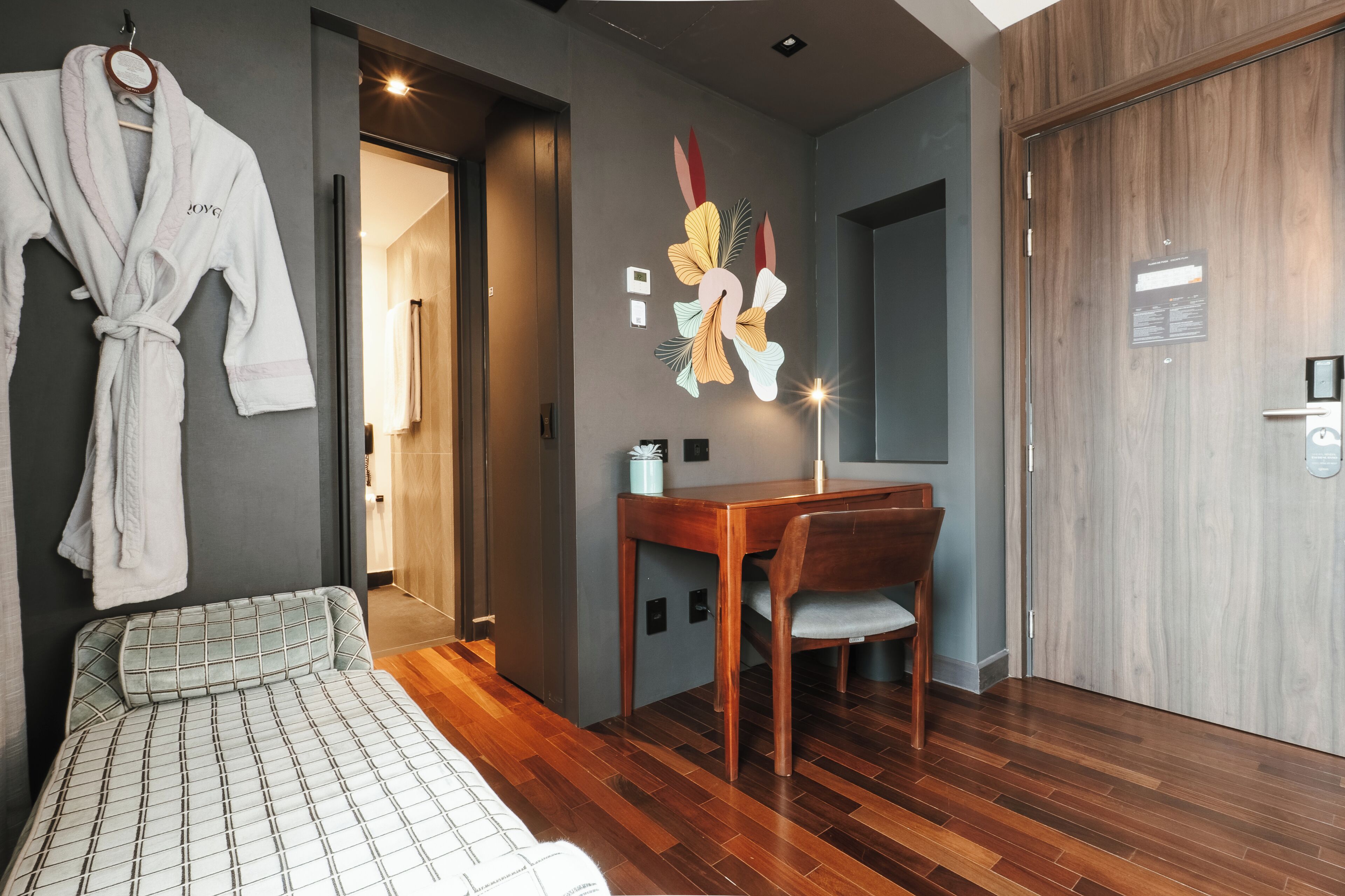 Una habitación de hotel moderna con piso de madera, una cama con colcha estampada, un escritorio con silla y una bata colgada en la pared.
