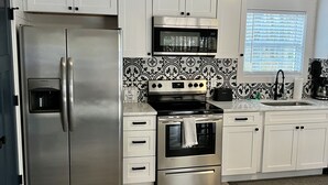 Una cocina moderna con armarios blancos, electrodomésticos de acero inoxidable, salpicadero blanco y negro y una ventana con persianas.