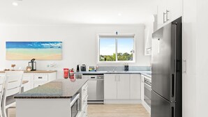 Et moderne køkken med hvide skabe, et rustfrit stål køleskab, et vindue med udsigt til grøn vegetation, og et maleri af en strand.