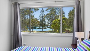 A bedroom with a large window, a bed with a striped bedspread, a lamp, and a view of trees and a lake.
