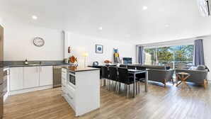 A modern kitchen with white cabinets, a dining area with a black table and chairs, and a living area with a grey sofa and a TV.