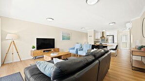 Ett modernt vardagsrum med en svart lädersoffa, ett träbord och en platt-TV. I bakgrunden finns en matplats med blå stolar och ett kök.