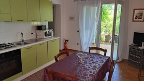 A kitchen with a gas stove, microwave, and sink. There is a dining table with chairs and a TV stand with a TV and storage shelves. - 3 Beautiful hillside holiday rental with sea views and pool (Nuoro)