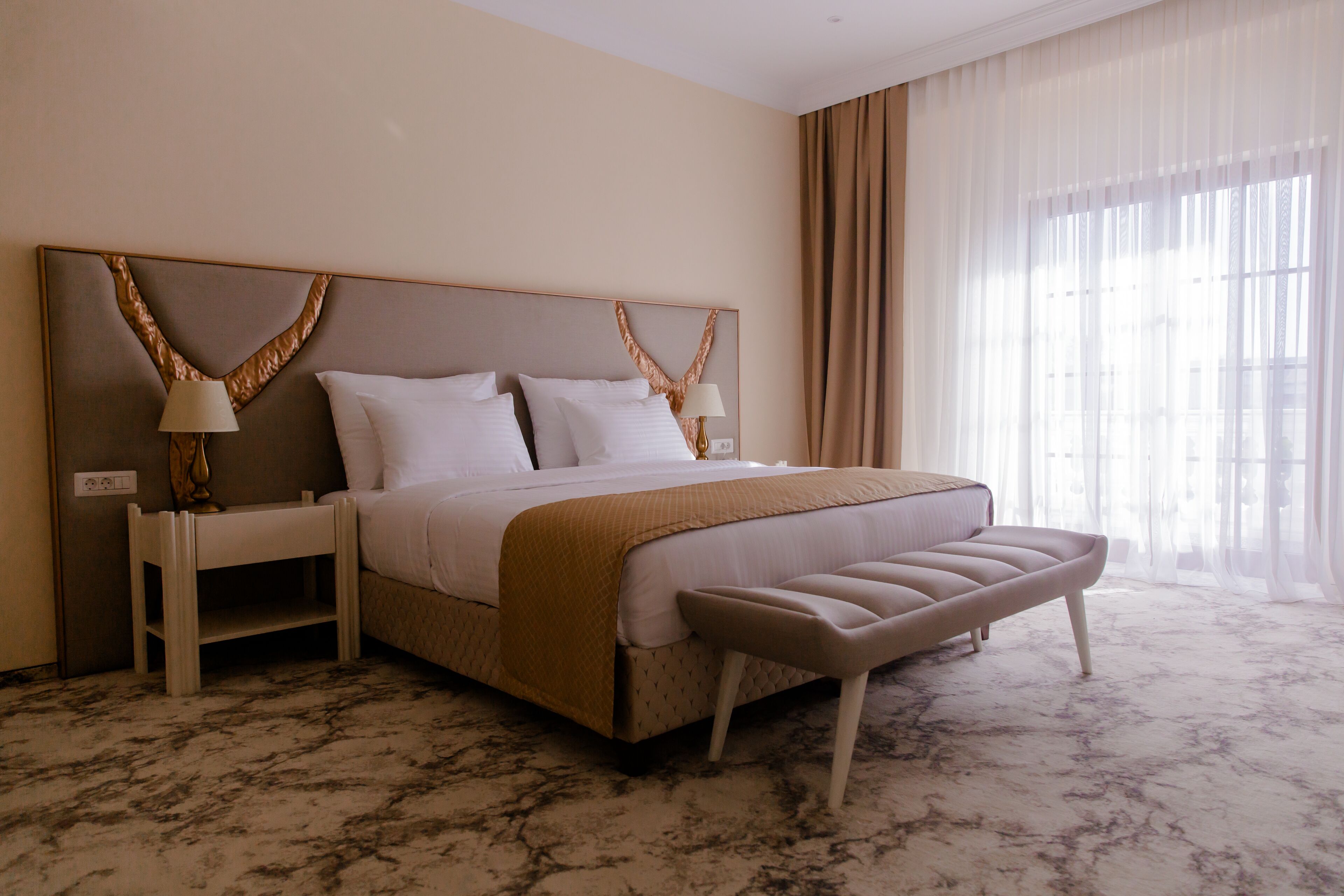 Habitación de hotel con una cama grande, dos mesitas de noche, un banco al pie de la cama y un ventanal con cortinas traslúcidas.