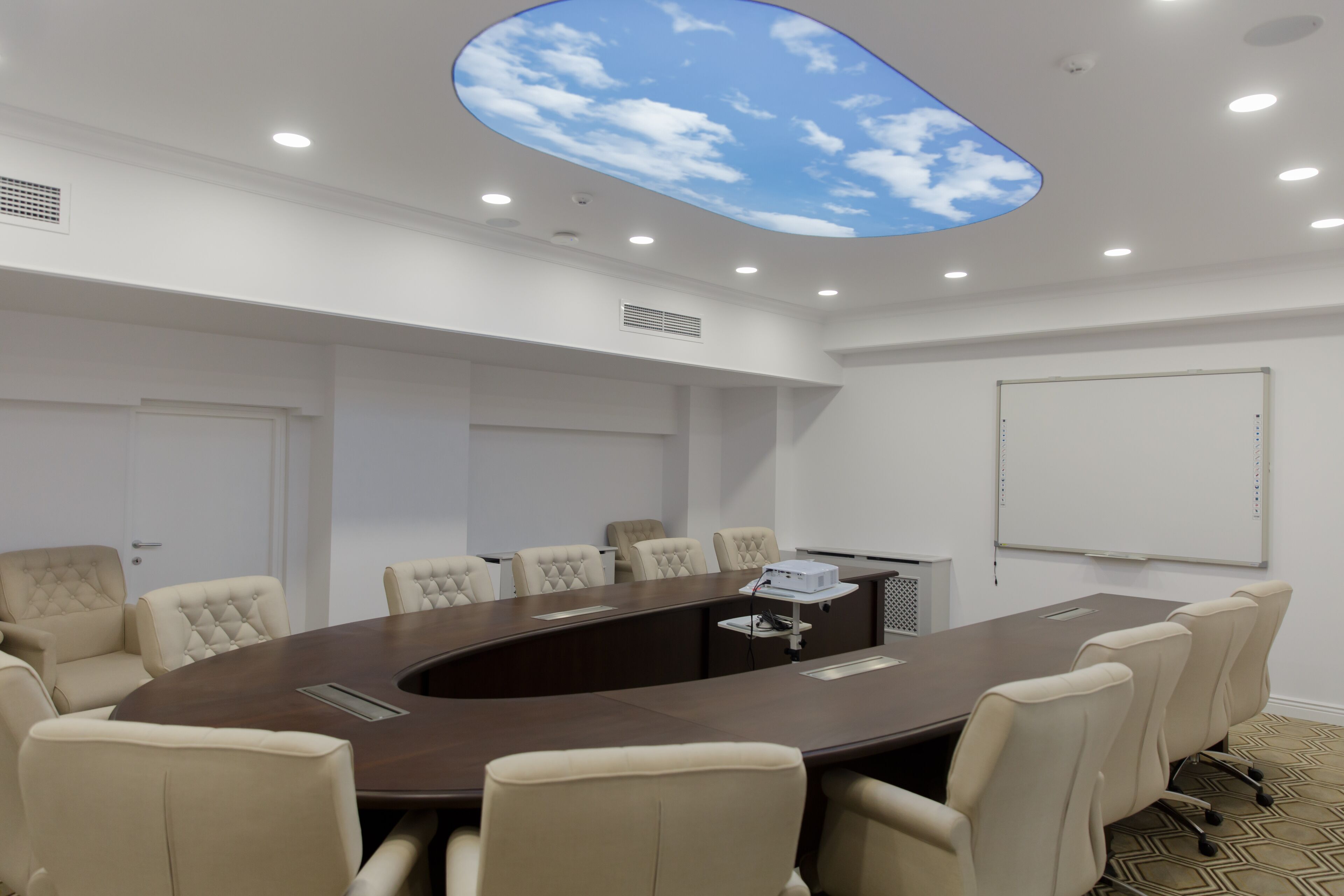 A conference room with a round table, beige chairs, a whiteboard, and a ceiling skylight.