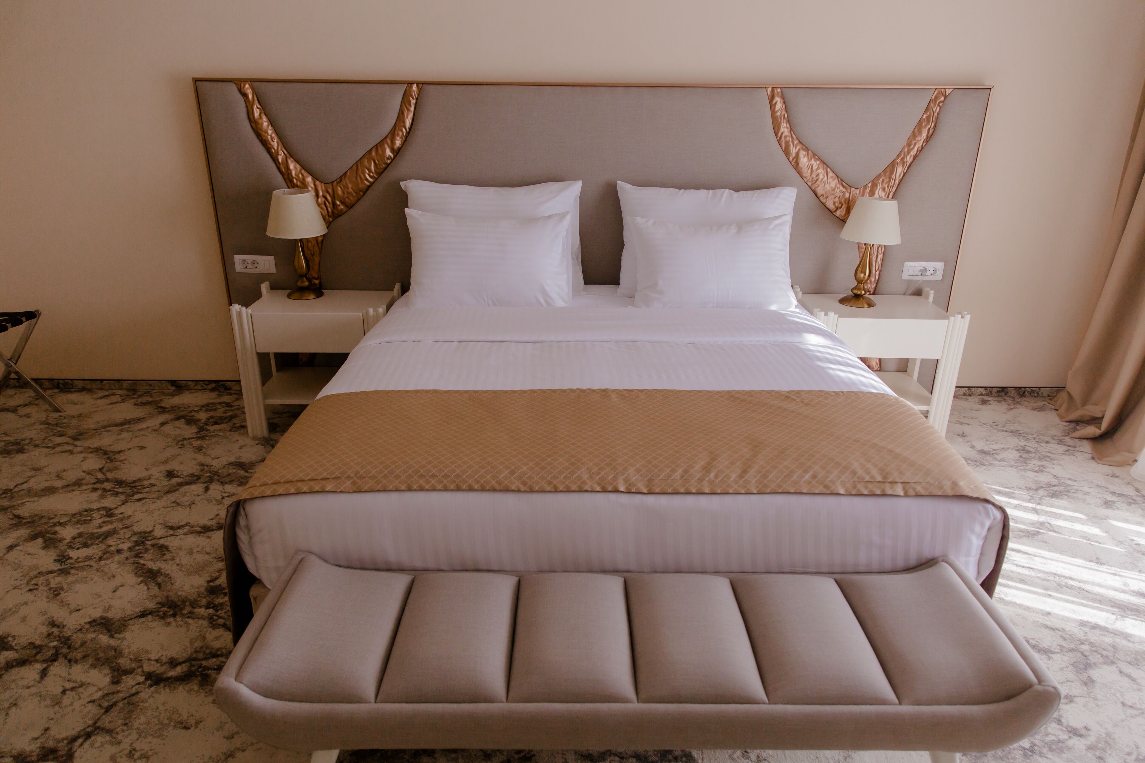 A hotel room with a large bed, two bedside tables with lamps, a headboard with a decorative design, and a marble floor.