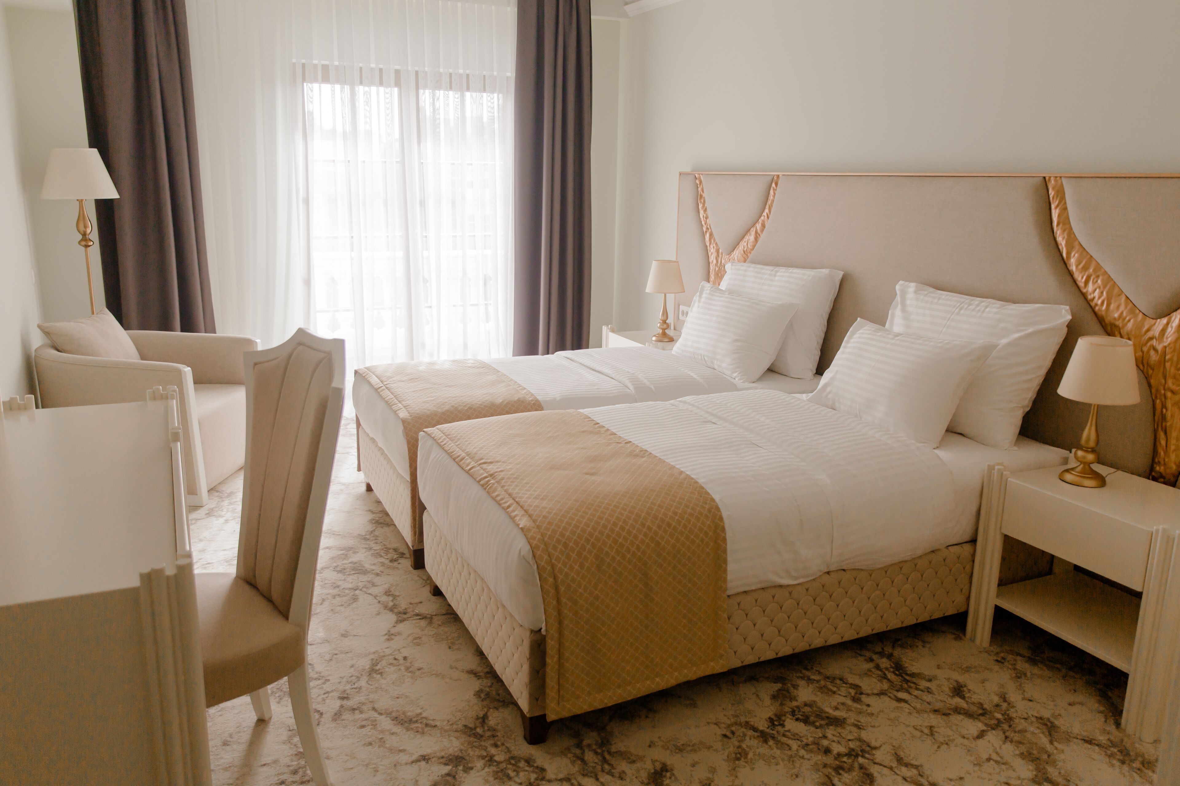 A hotel room with two beds, a sitting area with a chair, a nightstand with a lamp, and a large window with curtains.