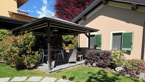 A covered outdoor seating area with a table and chairs, surrounded by greenery and a stone pathway.