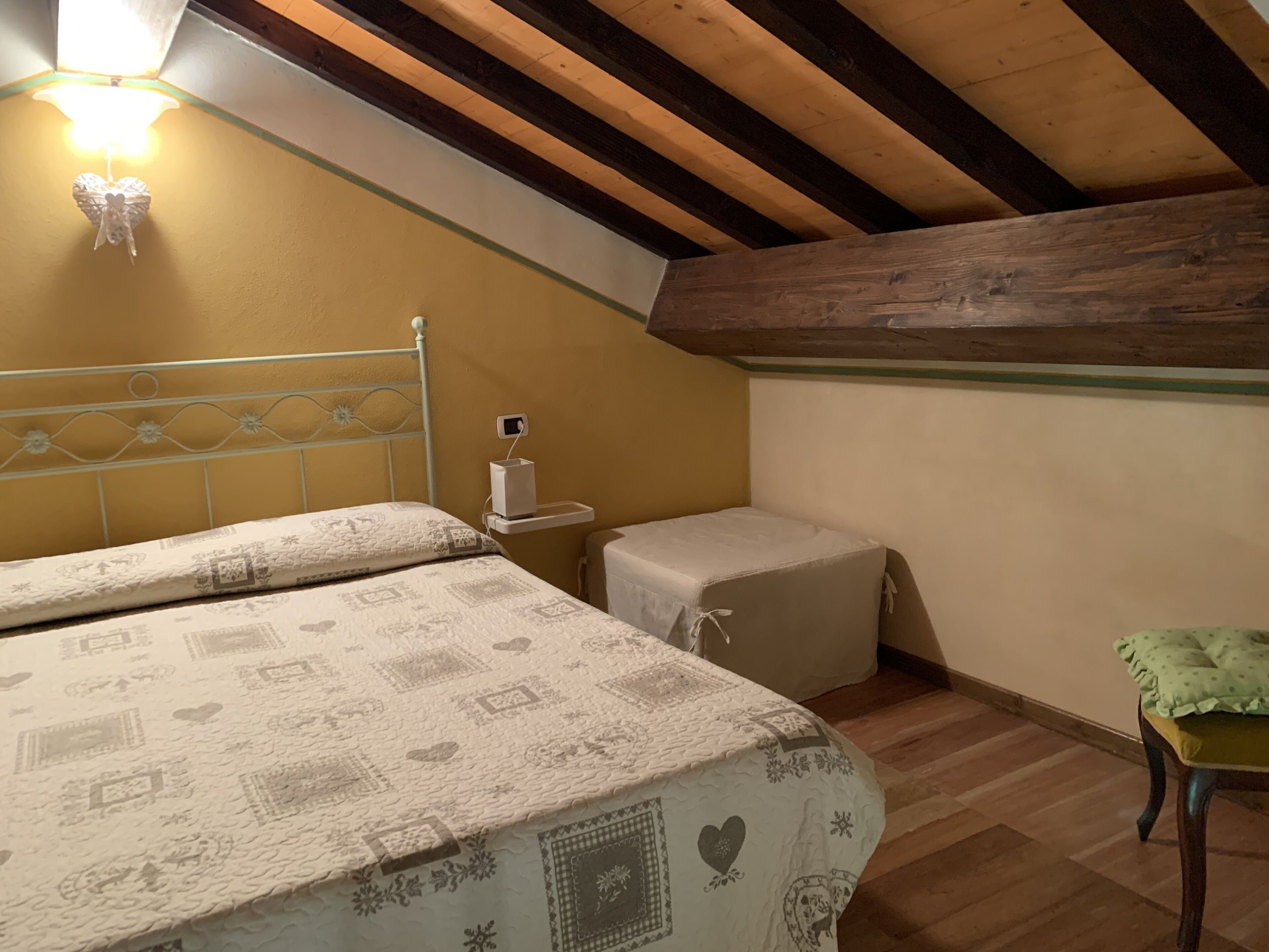 A small, cozy attic bedroom with a sloped ceiling, a bed with patterned bedding, a bedside table with a lamp, and a small stool.