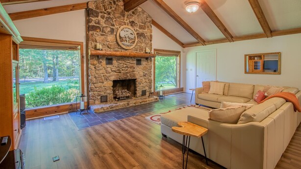 Un salon spacieux doté d’une cheminée en pierre, de poutres en bois, de grandes fenêtres et d’un canapé beige.